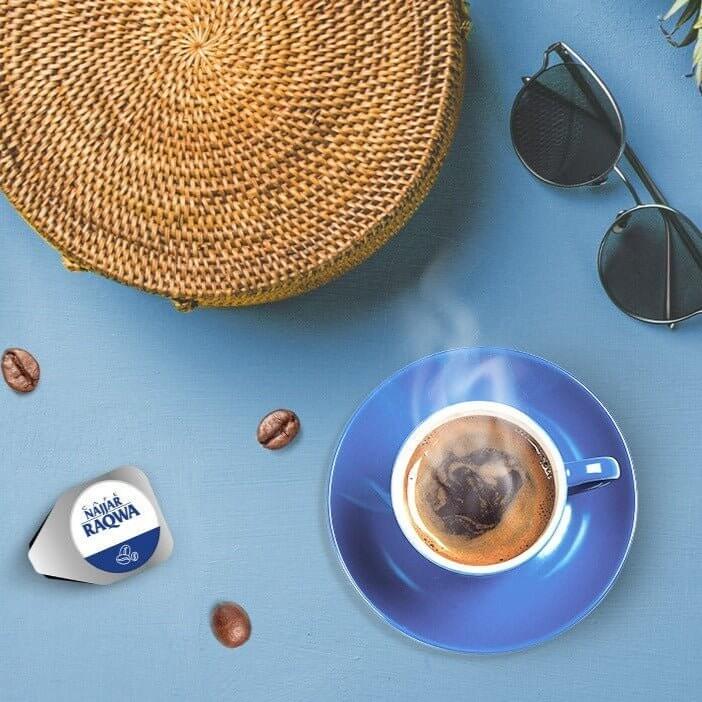 Najjar Raqwa Coffee Machine with steaming coffee cup, coffee beans, and sunglasses on a blue table.